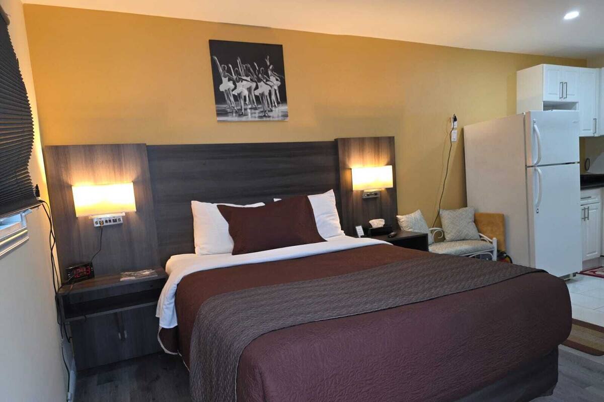 Quarto de hotel com uma cama grande, luminárias de cabeceira, uma mesinha, uma cadeira, um frigorífico e um quadro na parede.