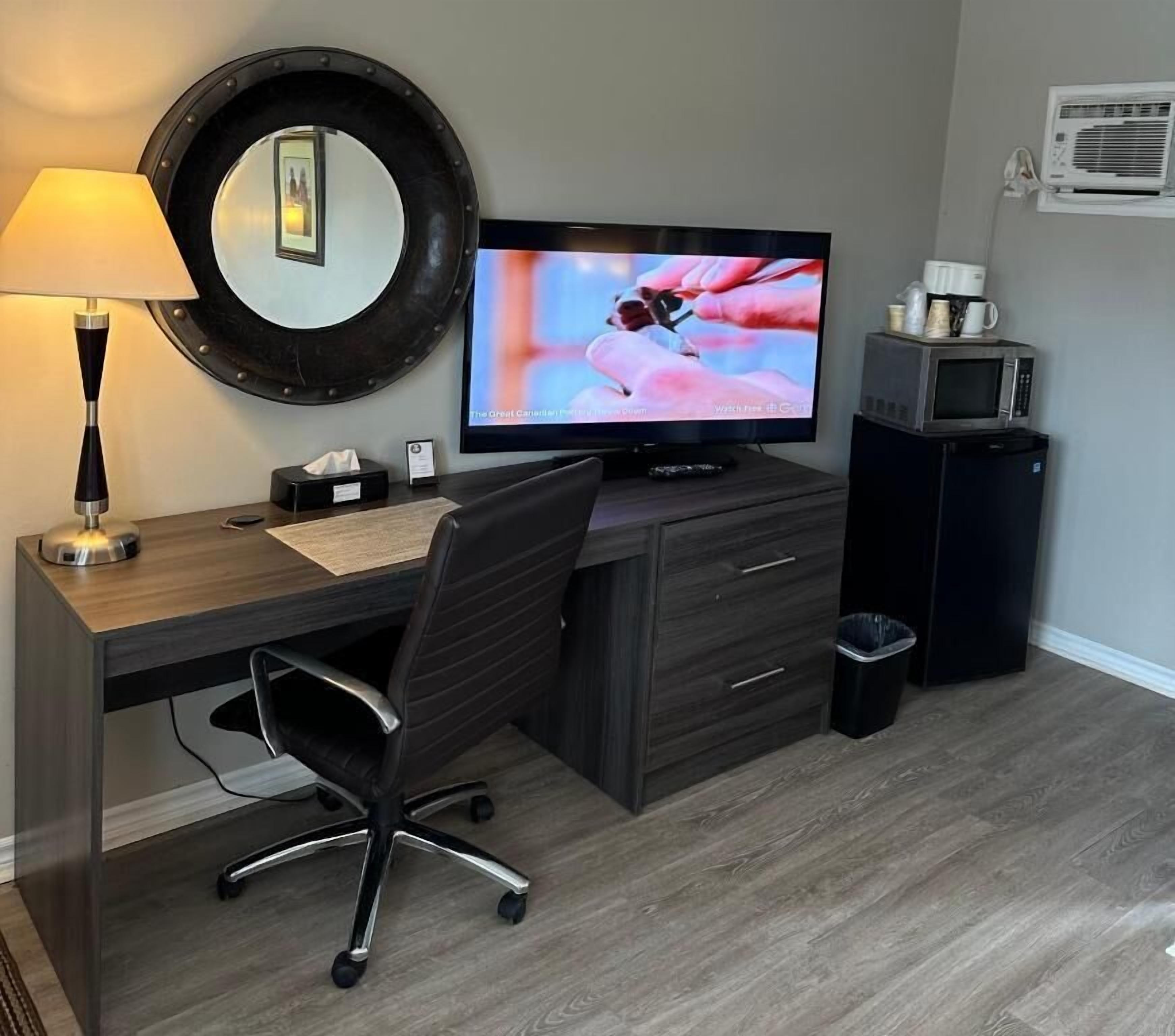 A hotel room with a desk, chair, television, lamp, and a trash bin.