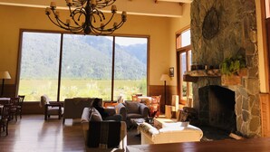 A spacious living room with a large window offering a mountain view, a stone fireplace, and a chandelier. - Hotel Natura Patagonia (Puerto Varas)
