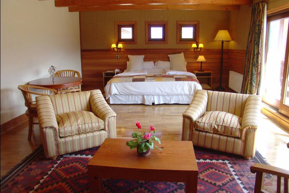 Ein gemütliches Hotelzimmer mit einem Bett, zwei Sesseln, einem kleinen Tisch mit Blumenvase und einem gemusterten Teppich.