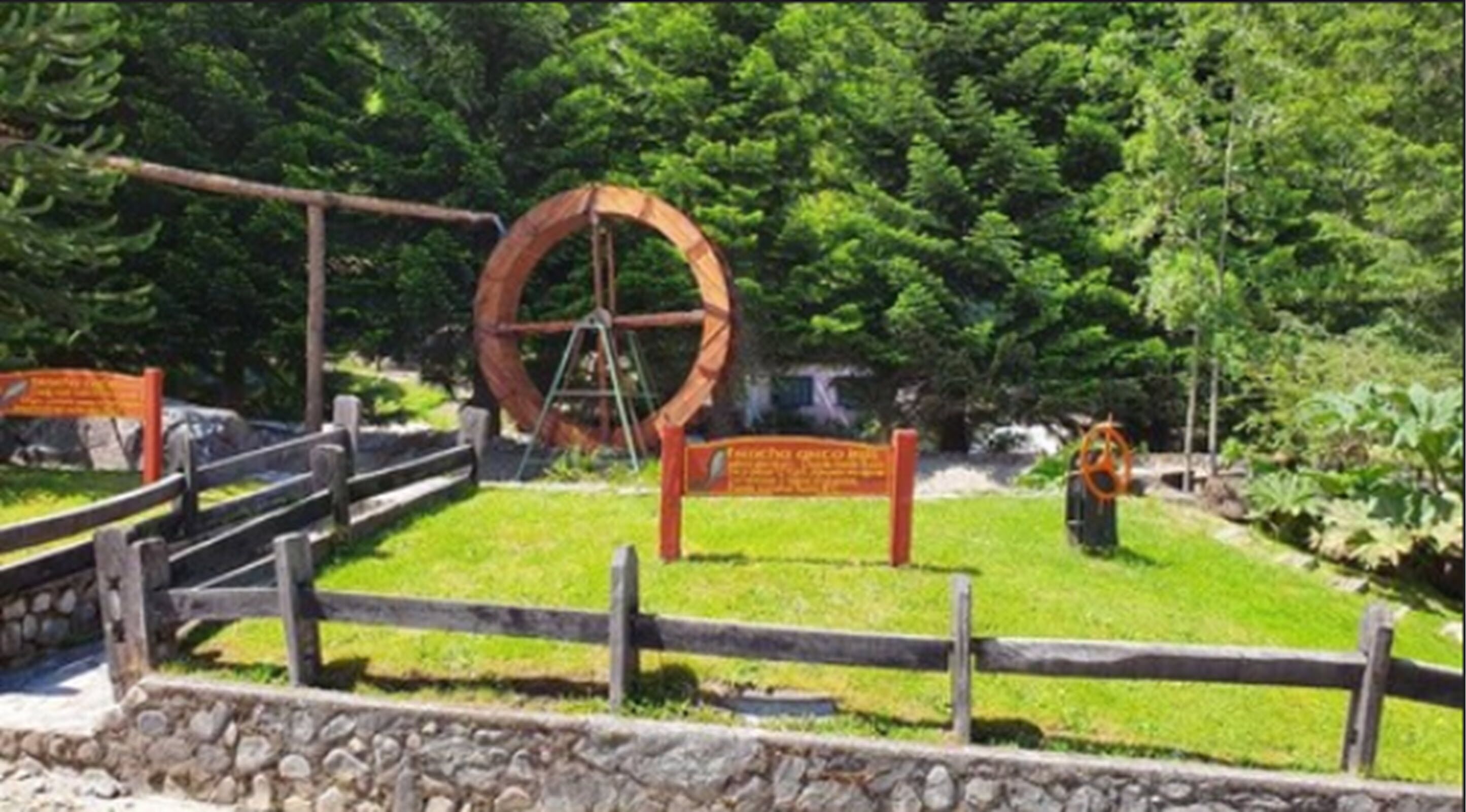 Una valla de madera, una estructura metálica circular y un cartel en un parque con abundante vegetación.