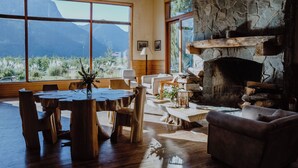 A spacious living room with a stone fireplace, wooden furniture, and large windows offering a mountain view. - Hotel Natura Patagonia (Puerto Varas)