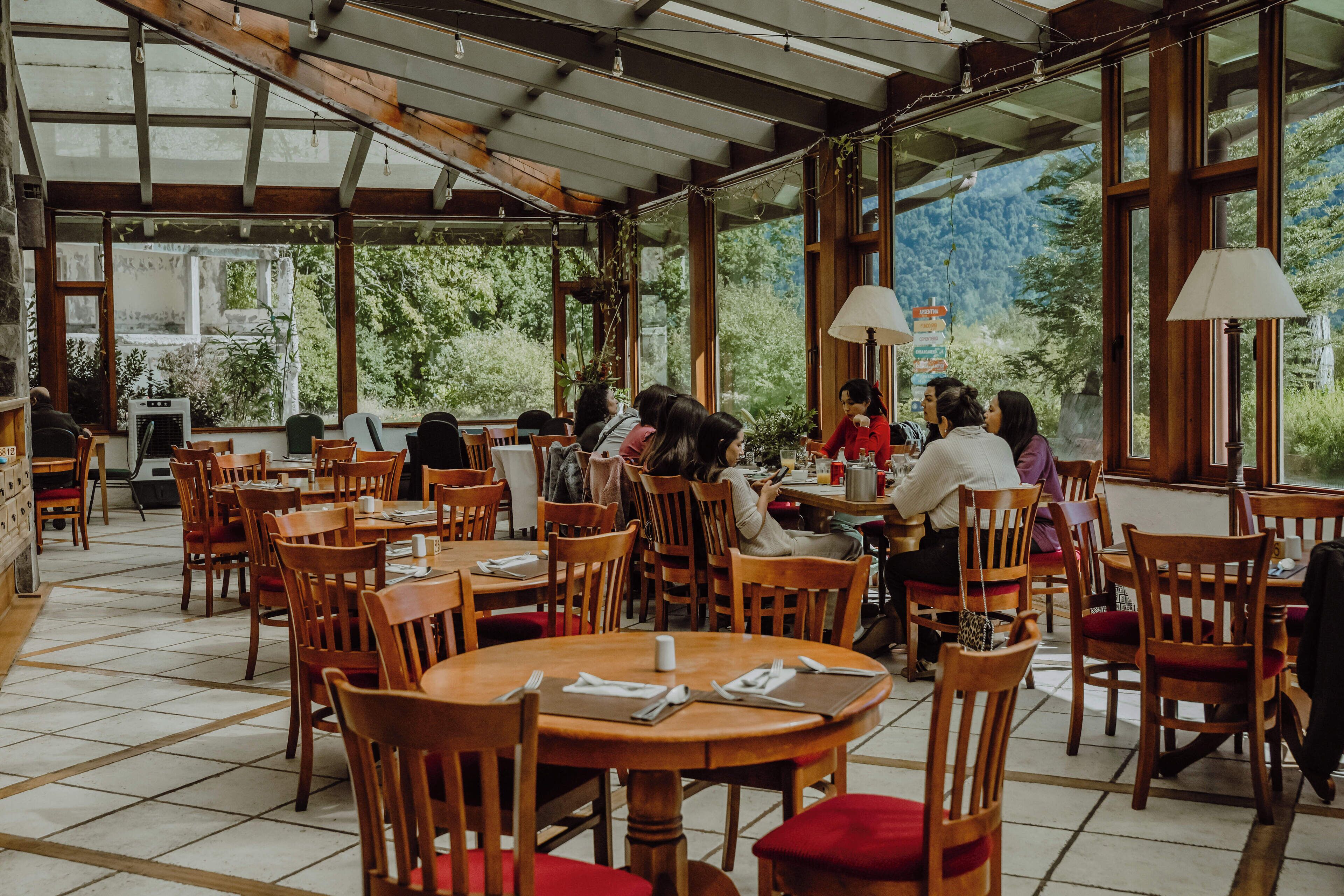 Un restaurante con mesas y sillas de madera, ventanas amplias y vistas a zonas verdes.