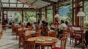 Un restaurante con mesas y sillas de madera, ventanas amplias y vistas a zonas verdes.
