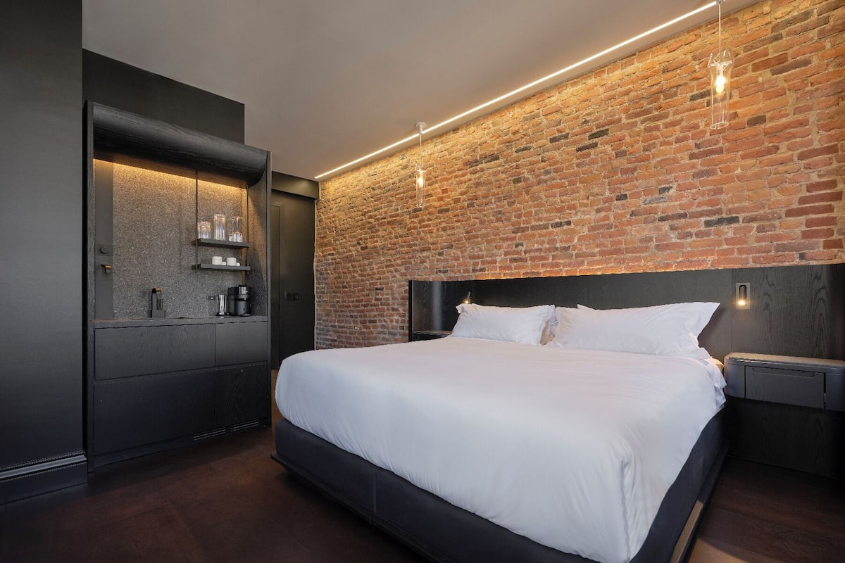 Ein modernes Schlafzimmer mit Backsteinwand, einem großen Bett, einer integrierten Kaffeemaschine und einer kleinen Ablagefläche mit Gläsern.