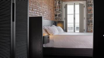 A modern bedroom with a brick wall, a large window, a bed with white linens, and a dark wooden wardrobe.