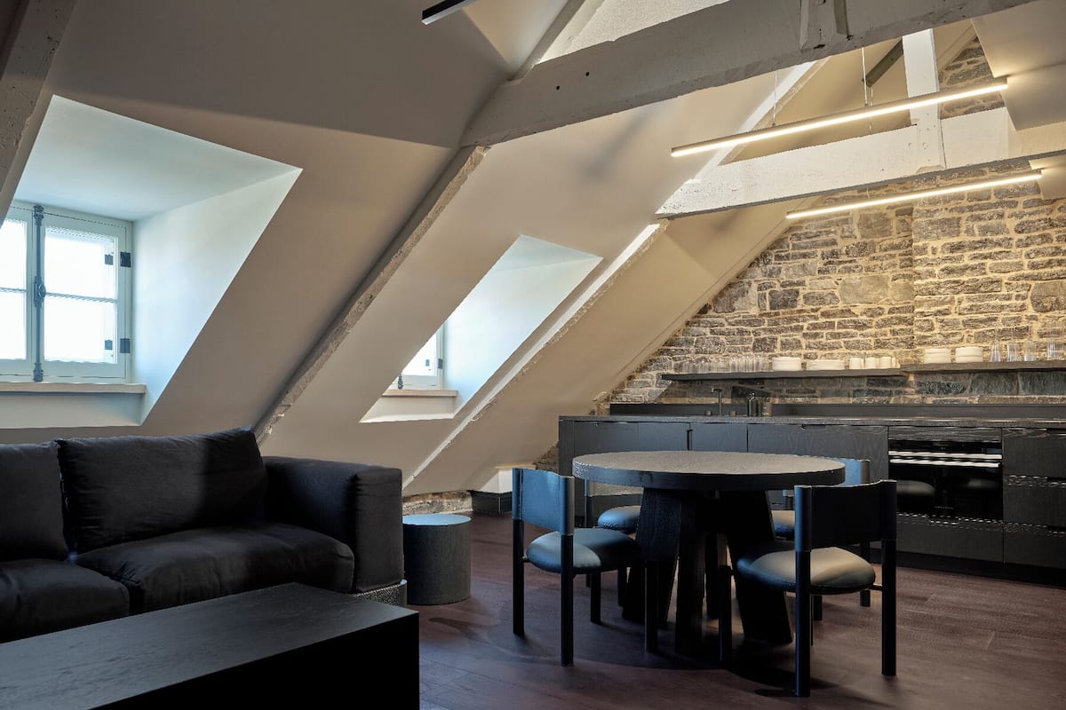 Ein modernes Loft mit schrägverdachter Decke, freiliegender Backsteinwand und einer gemütlichen Sitzecke mit dunkler Couch und rundem Tisch.