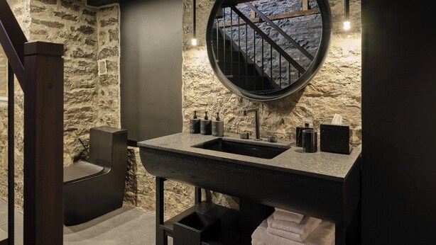 A bathroom with a stone wall, a round mirror, a sink with a black countertop, and a towel rack. - Hôtel Maison du Fort (Québec City)