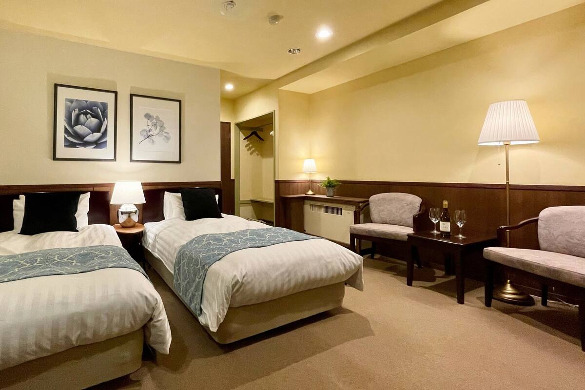 A hotel room with two beds, a desk, a chair, a lamp, and two framed pictures on the wall.