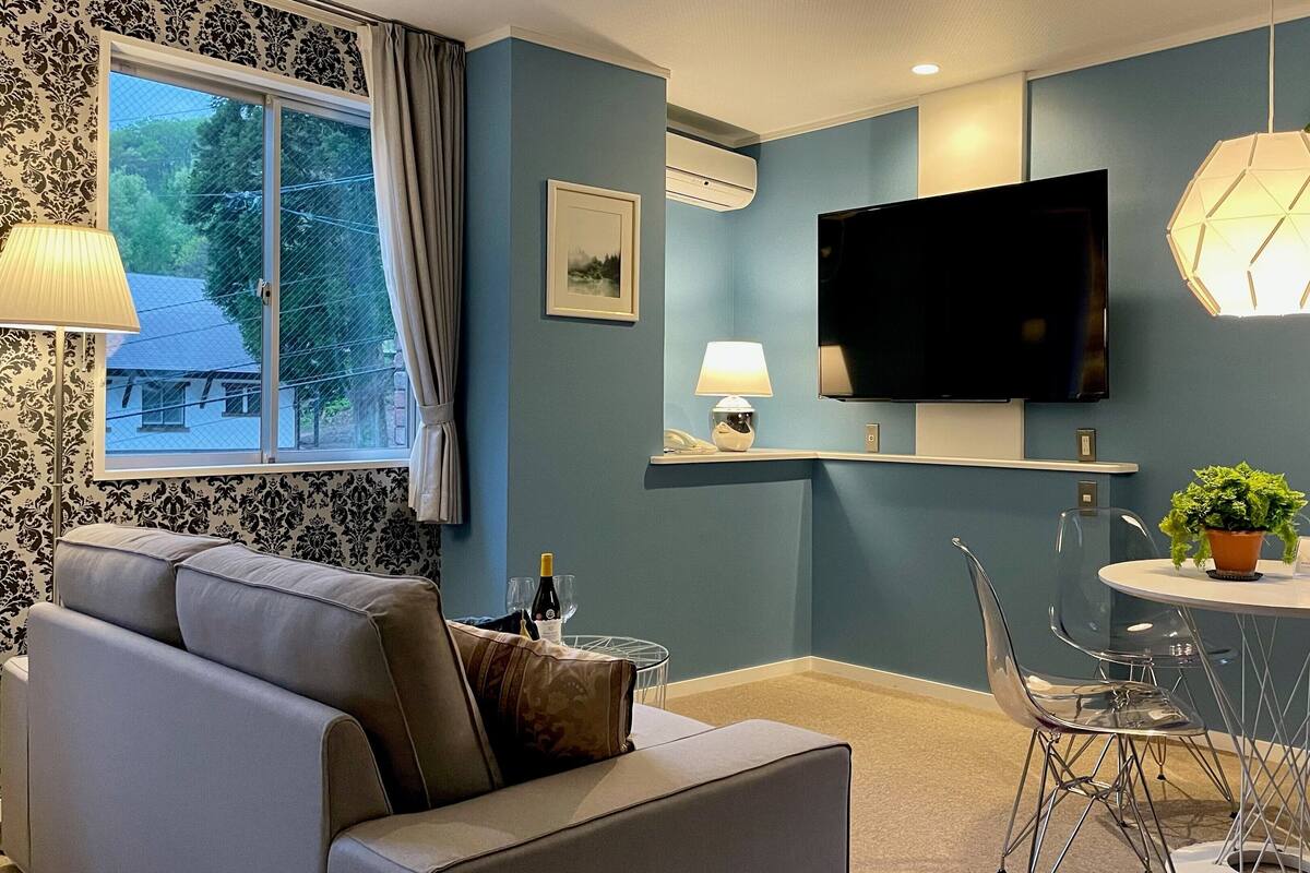 A modern living room with a grey sofa, a dining table with chairs, a flat-screen TV mounted on the wall, and a potted plant.