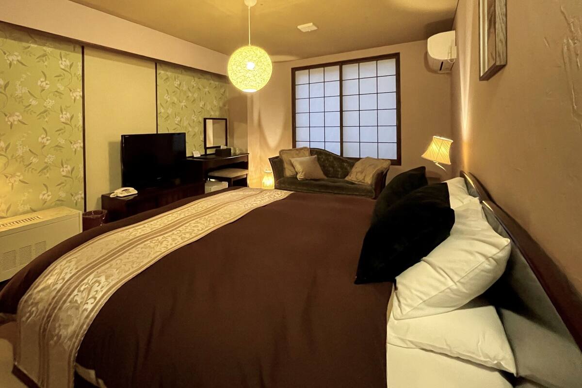A bedroom with a bed, a desk, a chair, a television, and a window.