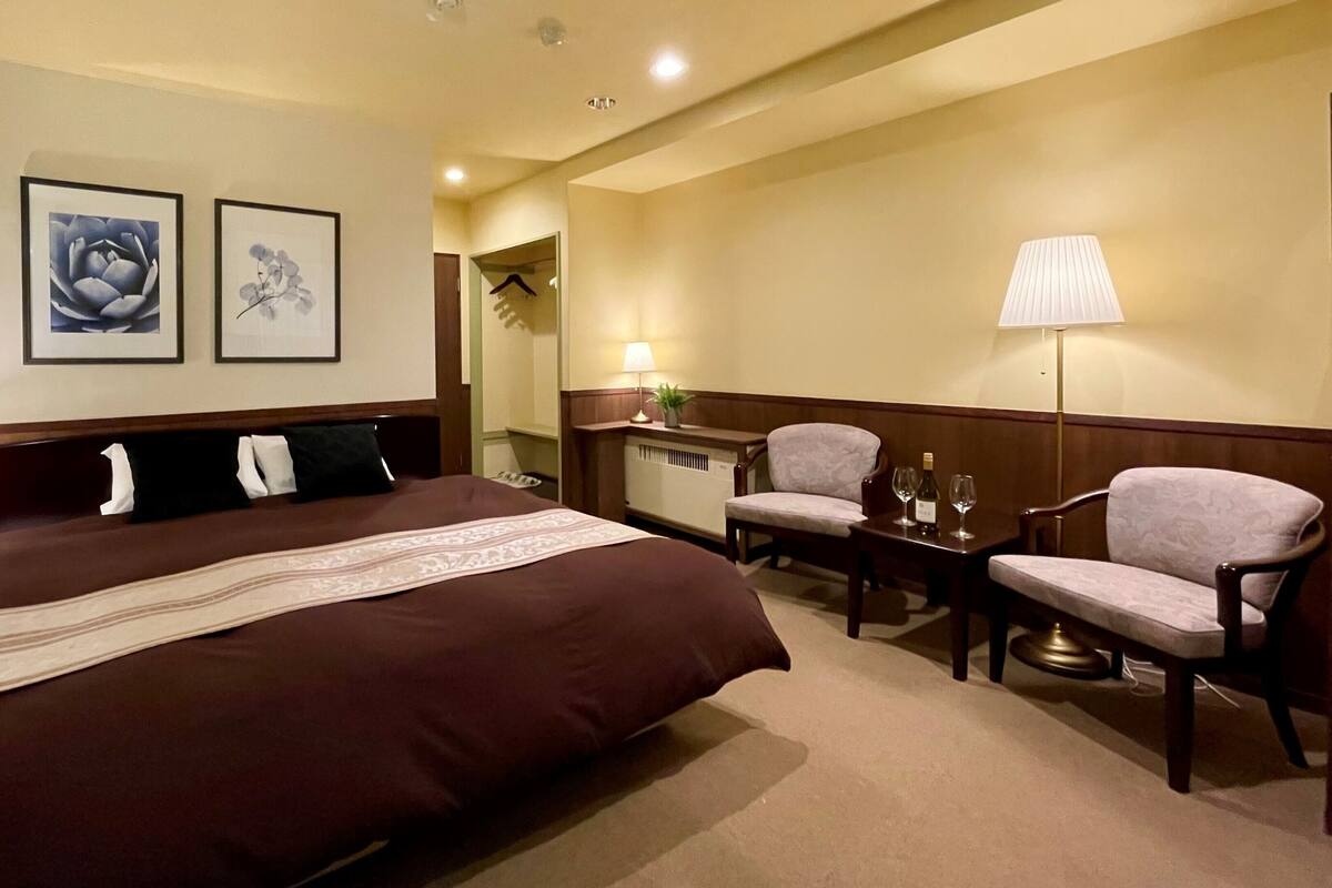 A hotel room with a large bed, two chairs, a desk with a lamp, and two framed pictures on the wall.