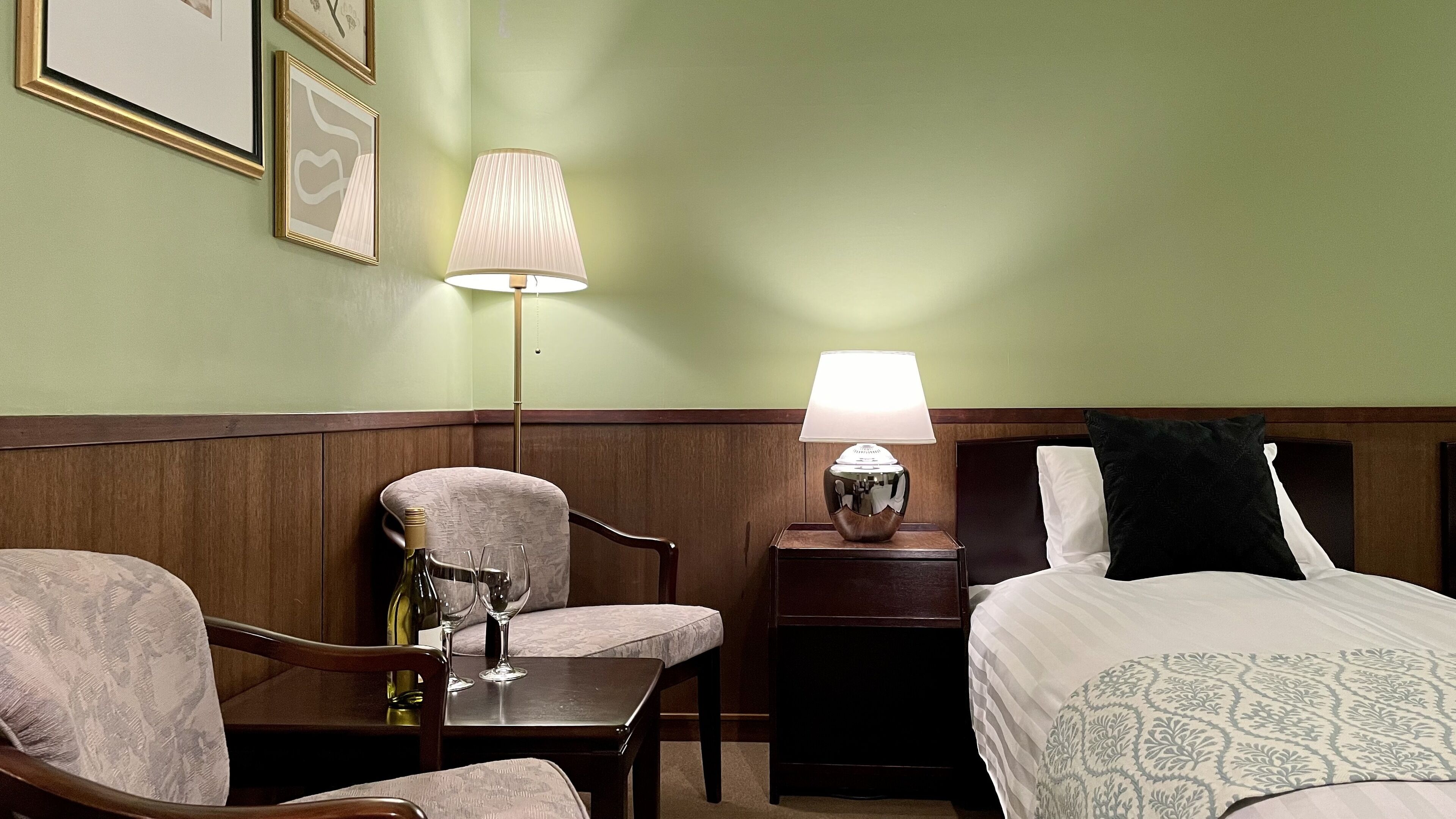 A hotel room with a bed, two chairs, a table with a bottle and glasses, and two lamps.