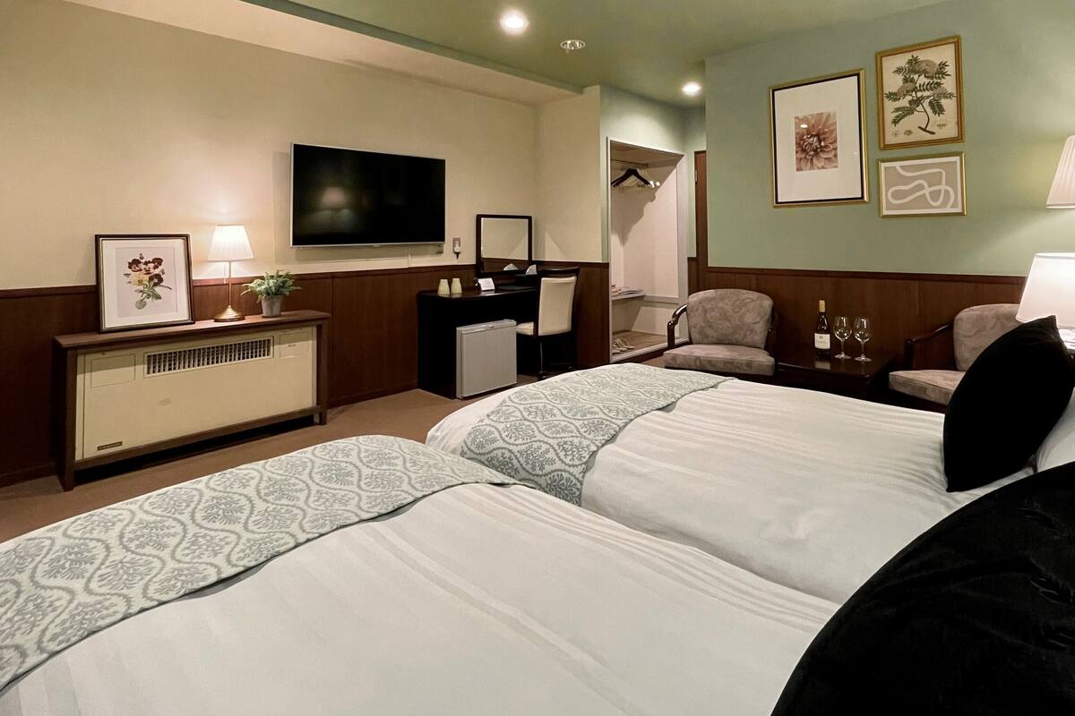 A hotel room with two beds, a desk, a TV, a chair, a lamp, and framed pictures on the wall.
