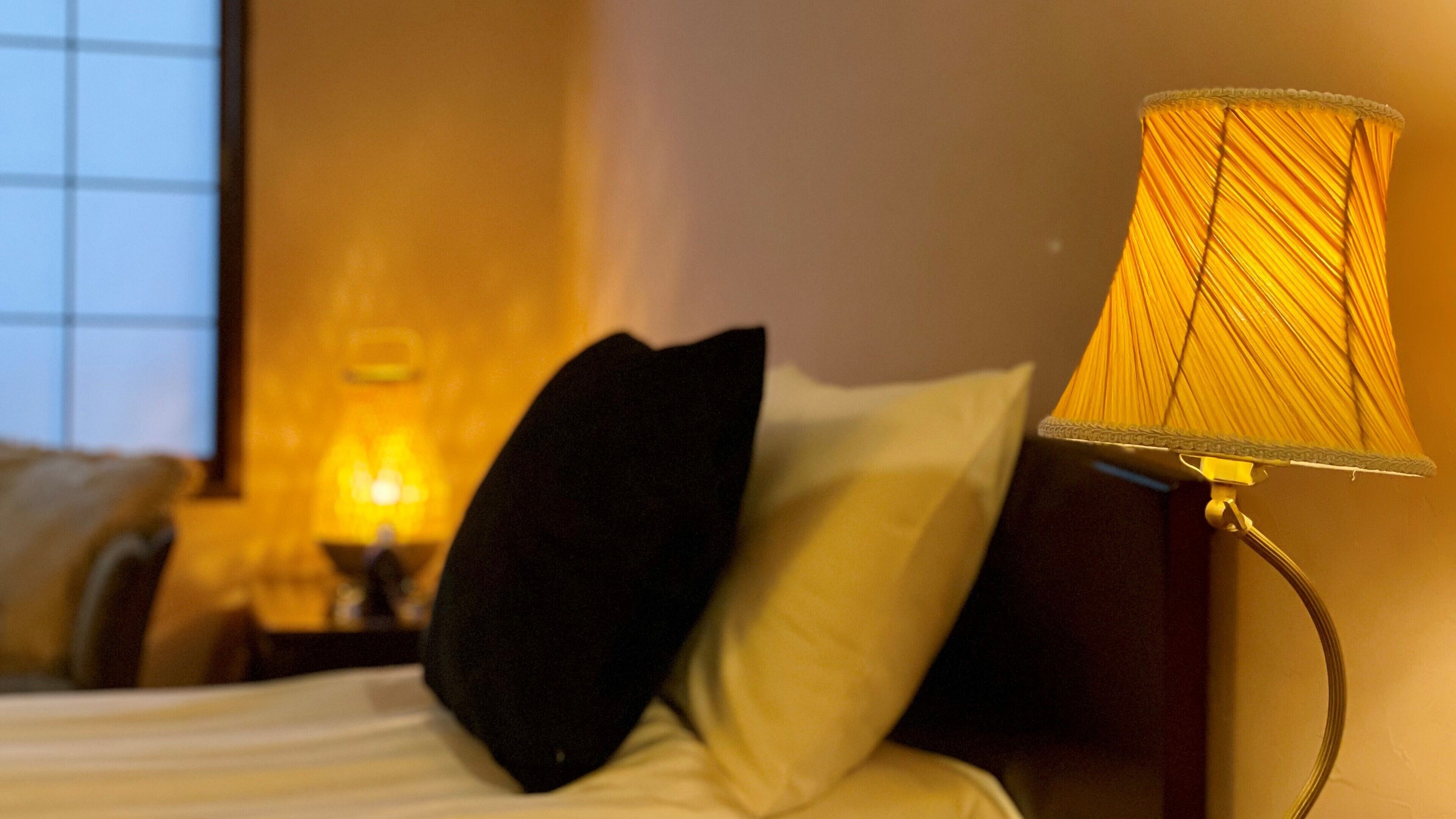 A bed with white linens, a black pillow, and a lamp with a yellow shade.