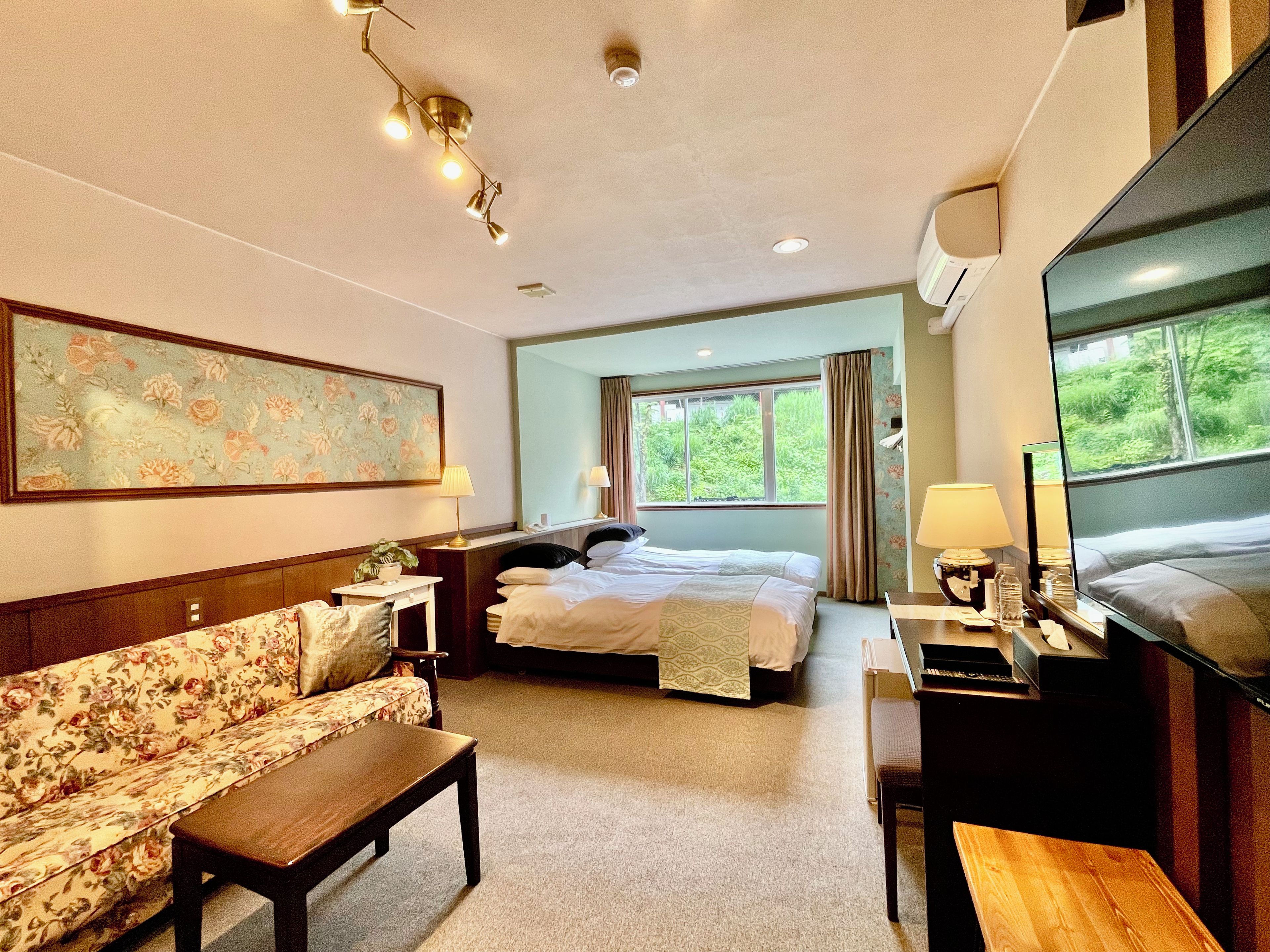 Quarto de hotel com cama, sofá, escrivaninha, televisão e janela com vista para o verde.