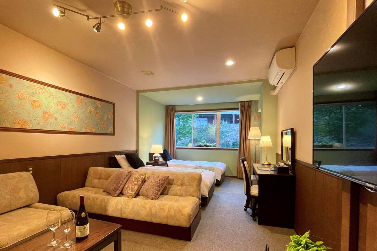 A hotel room with a sofa, a bed, a desk with a lamp, a mirror, and a window with a view of trees.