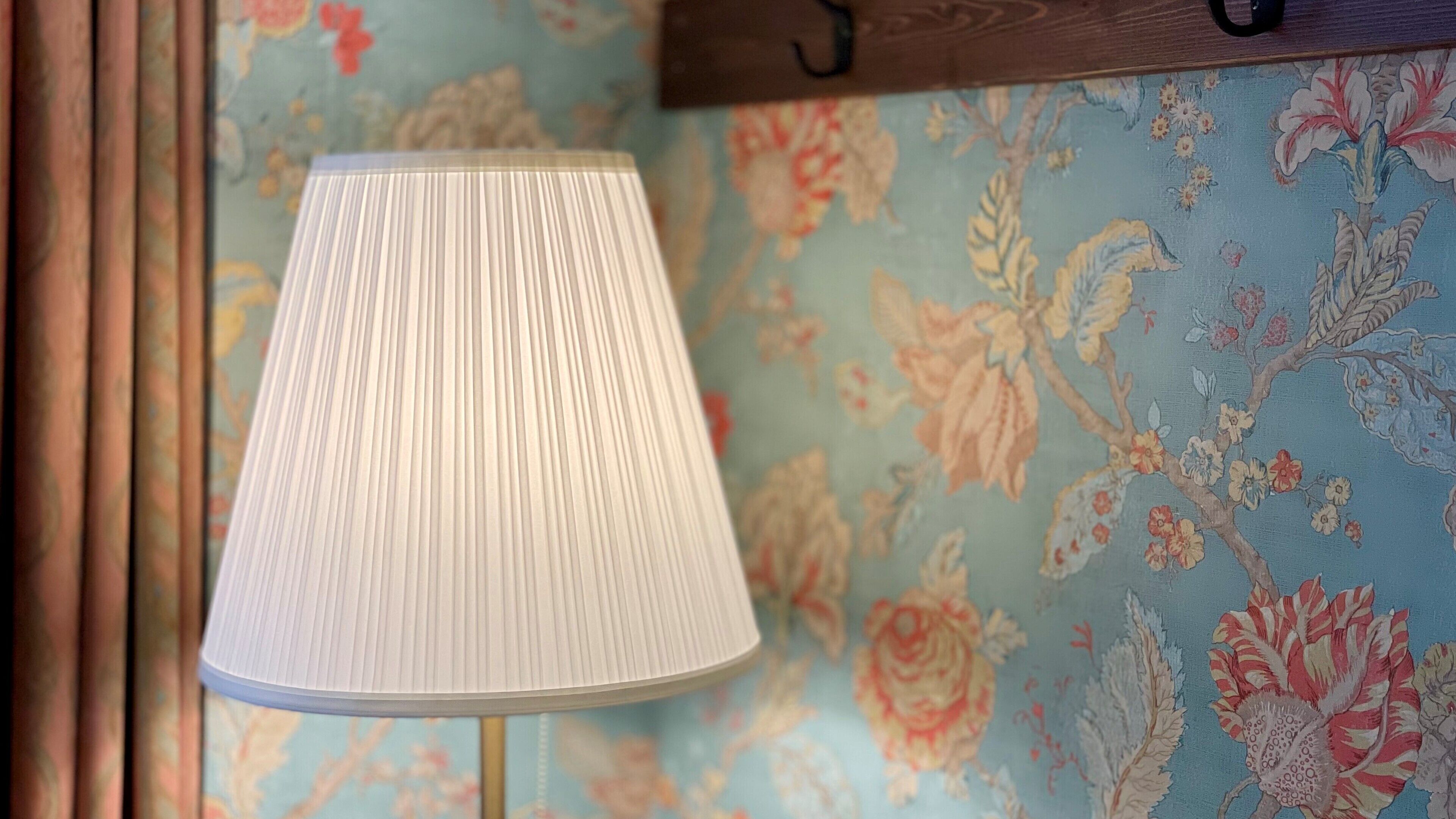 A lamp with a ribbed lampshade on a table against a floral wallpaper background.