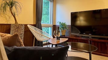 Uma sala de estar com uma televisão de tela plana grande, uma poltrona de vime, uma mesa de vidro com taças de vinho e um vaso de planta.