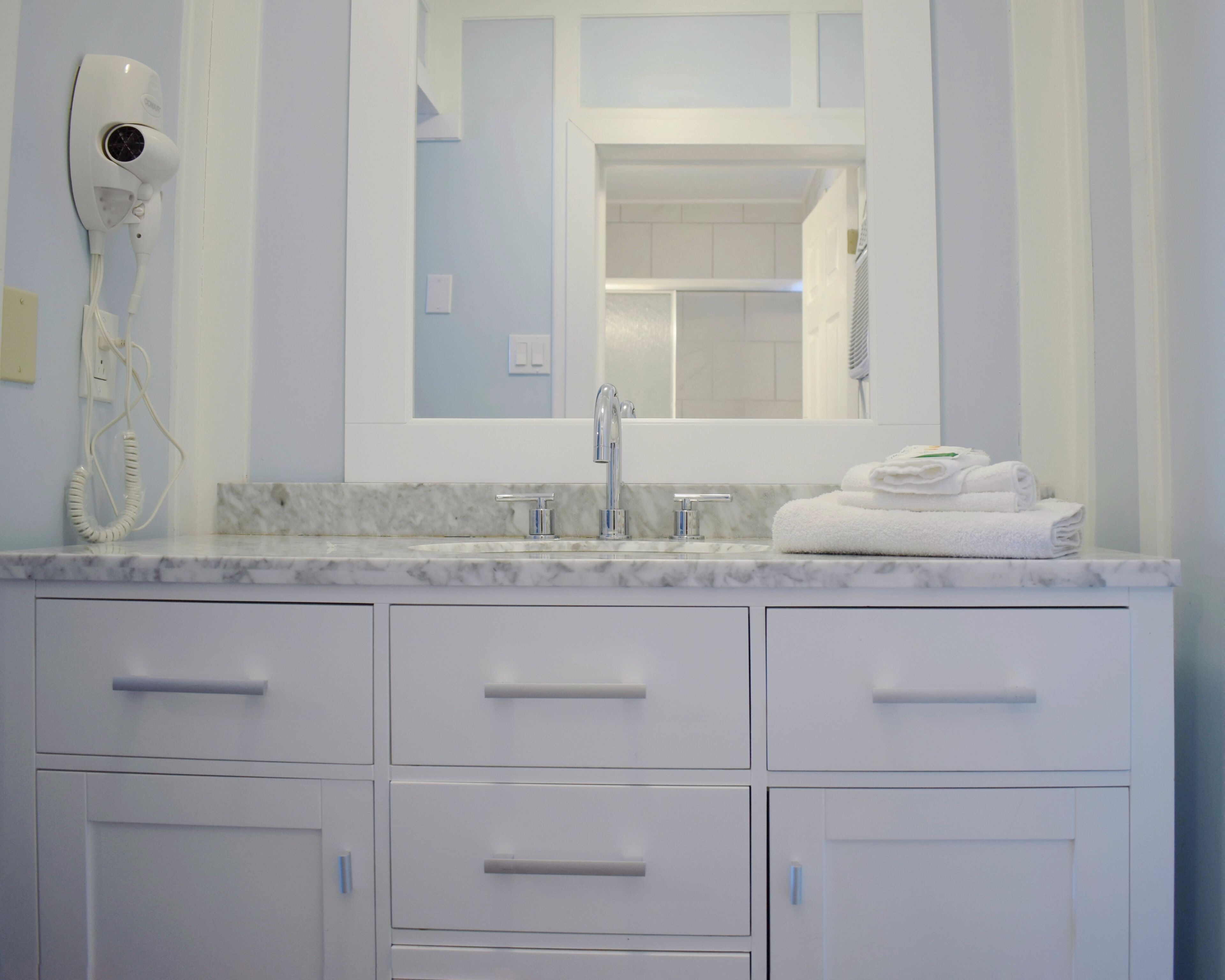 Une salle de bain dotée d’un comptoir en marbre, d’un lavabo avec un robinet moderne, d’un miroir au-dessus du lavabo et d’un sèche-cheveux fixé au mur.
