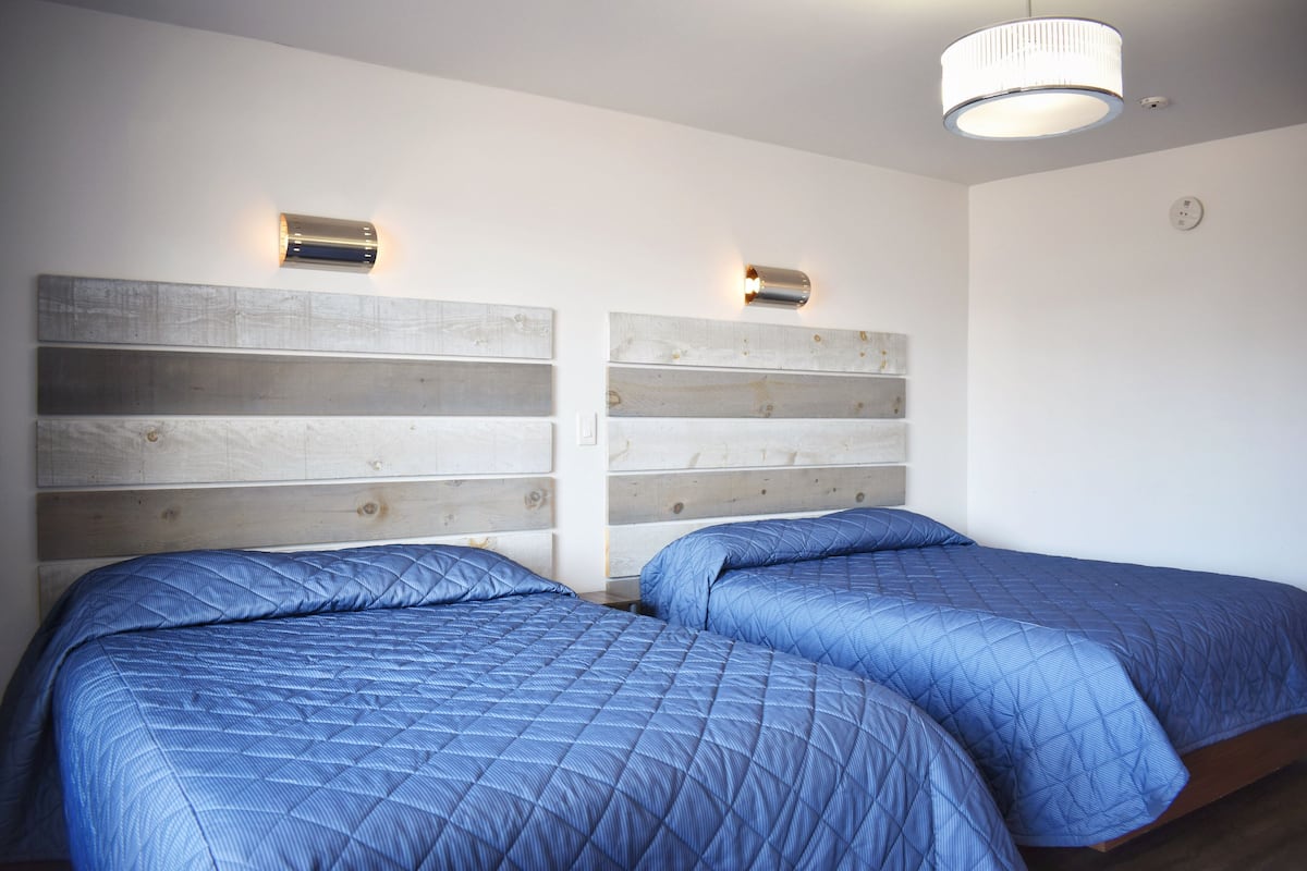 Una habitación con dos camas, cada una con cubrecama azul acolchado y cabecero de madera. Hay una lámpara en el techo sobre las camas.