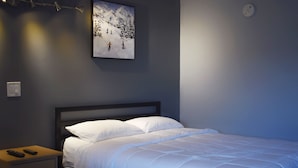 A bedroom with a bed, a nightstand, a remote control, and a painting of skiers on a mountain. - Elect Inn 5 (Cornwall)