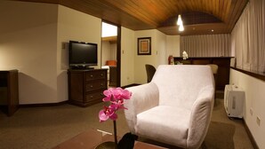 A room with a wooden ceiling, a television on a cabinet, a white armchair, and a small table with a pink orchid. - J&A Classic Hotel (Lima)