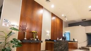 A modern reception desk with a wooden finish and a marble counter, a sign indicating a maximum occupancy of 100 people, and a potted plant with yellow flowers. - J&A Classic Hotel (Lima)