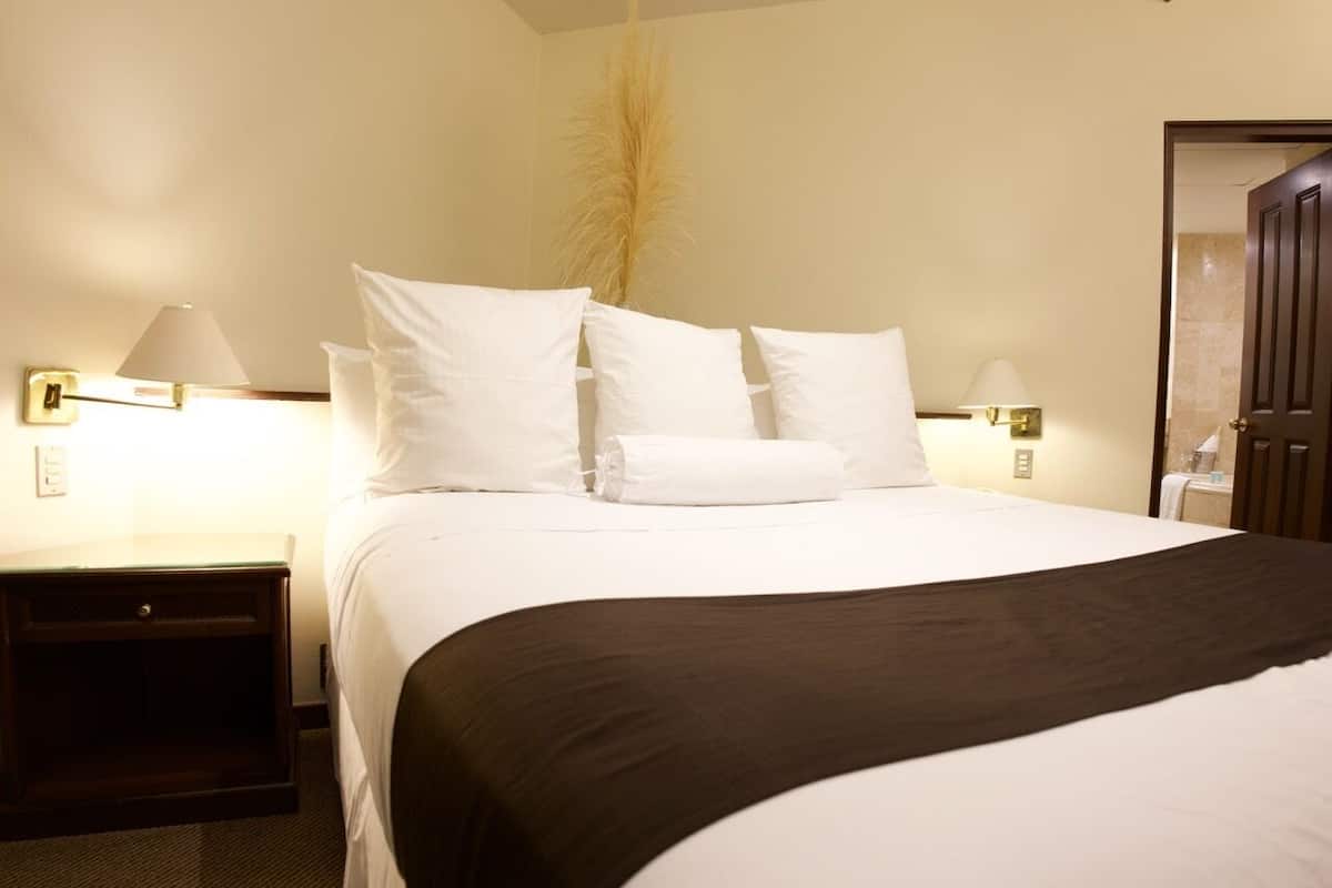 Uma cama bem arrumada com lençóis brancos e um cobertor escuro, com duas mesas de cabeceira com abajures, em um quarto com uma porta e uma luminária fixada na parede.