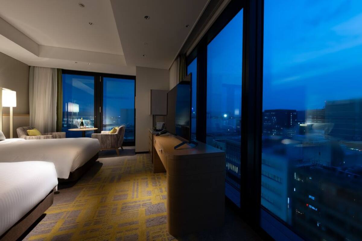 Une chambre d’hôtel avec deux lits, un bureau, une chaise et une grande fenêtre donnant sur la ville.