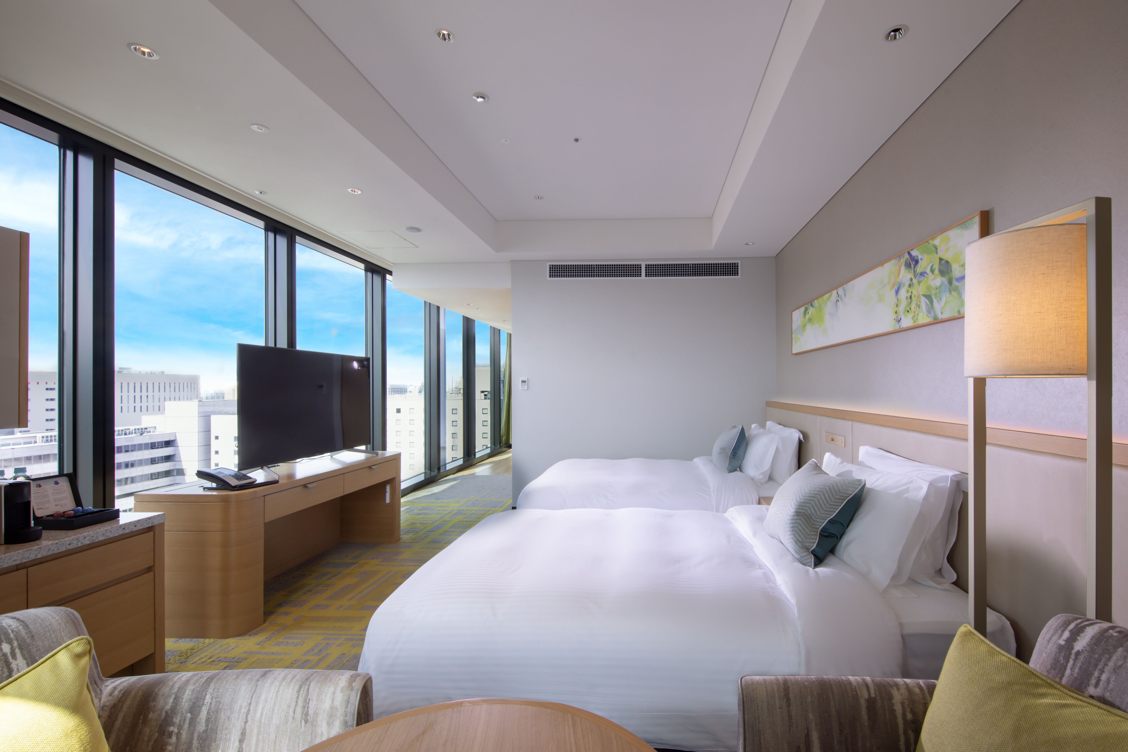 A hotel room with two beds, a desk with a flat-screen TV, a large window with a city view, and a painting on the wall.