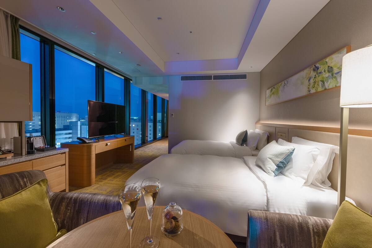 Une chambre d’hôtel dotée d’un grand lit, d’un bureau avec une télévision, d’une table à manger avec des verres, et offrant une vue sur la ville grâce à de grandes fenêtres.