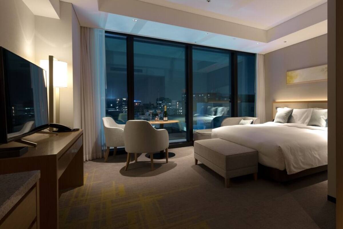 Una moderna camera d'albergo con un letto grande, una scrivania con una lampada, una sedia e vista sulla città illuminata.