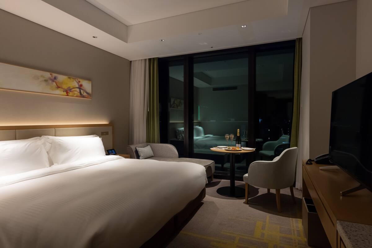 Une chambre d’hôtel moderne dotée d’un grand lit, d’un bureau avec un téléviseur à écran plat, d’une petite table ronde avec une bouteille et des verres, et offrant une vue sur la ville grâce à de grandes fenêtres.