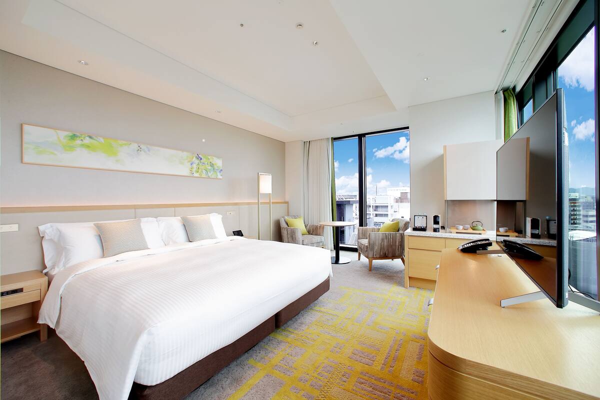 Une chambre d’hôtel moderne dotée d’un grand lit, d’un bureau avec un ordinateur, d’une chaise, d’une petite table et d’une grande fenêtre donnant sur la ville.