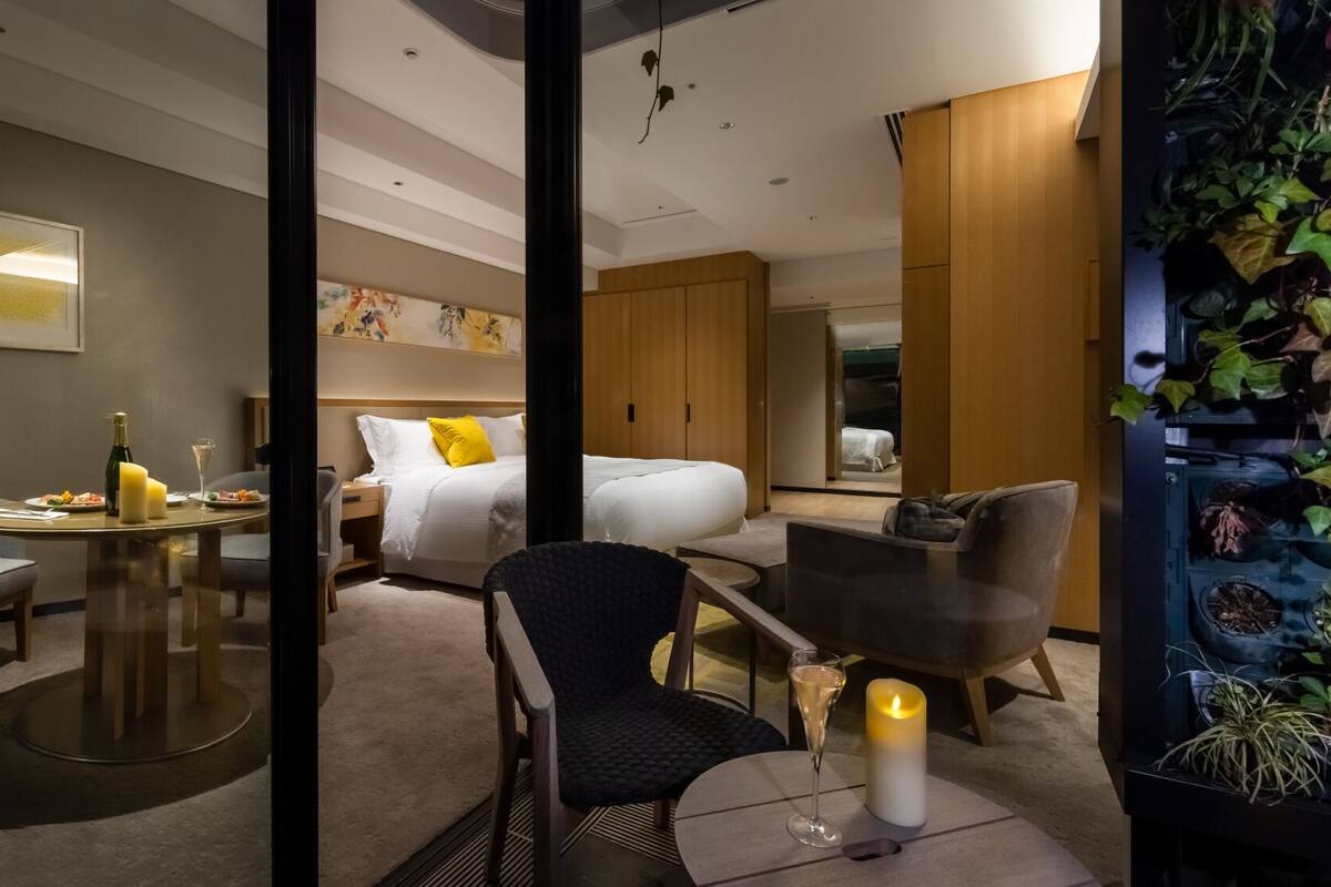 Une chambre d’hôtel comprenant un lit, une table à manger avec des chaises, un petit coin salon et un présentoir à plantes fixé au mur.