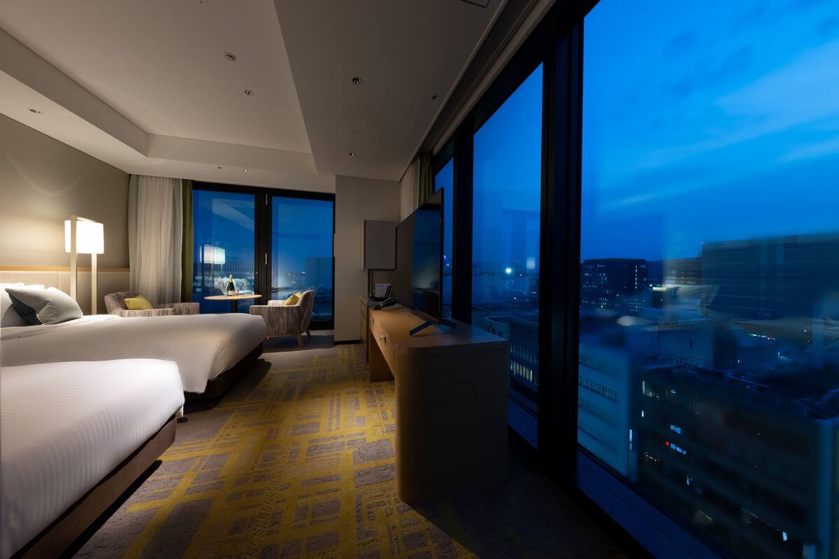 Une chambre d’hôtel avec deux lits, un bureau, une chaise et une grande fenêtre donnant sur la ville.