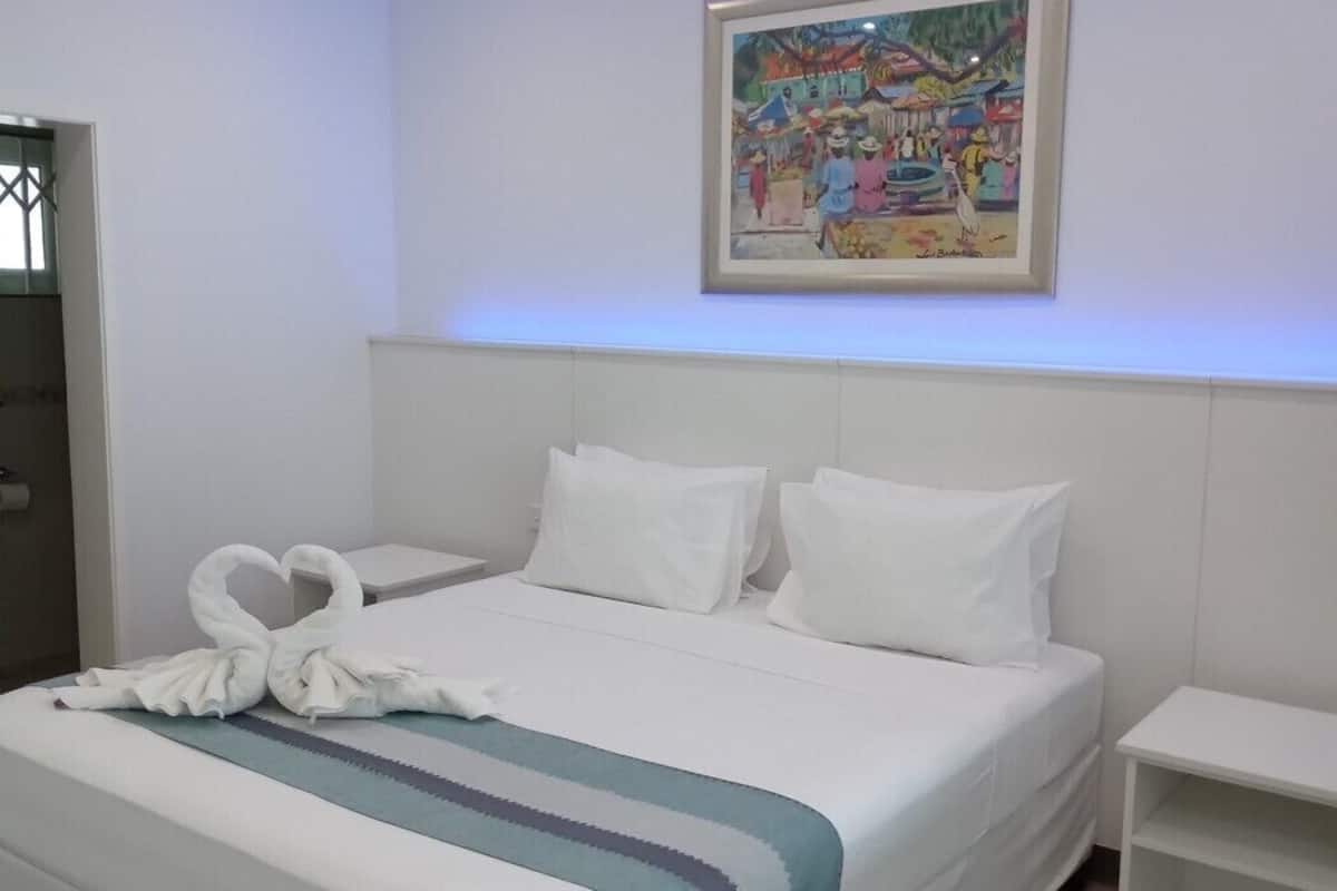 A hotel room with a bed, white pillows, a blue and white striped blanket, a white nightstand, a framed painting on the wall, and a ceiling fan.