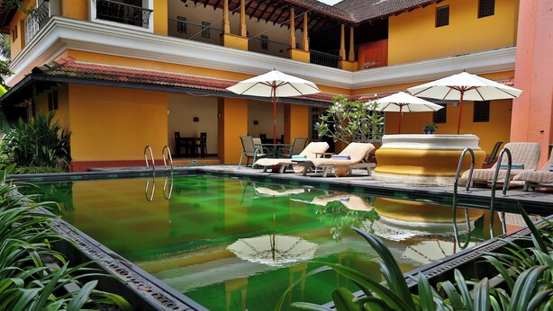 Une piscine entourée de plantes et d’un bâtiment jaune, avec des chaises longues et des parasols.
