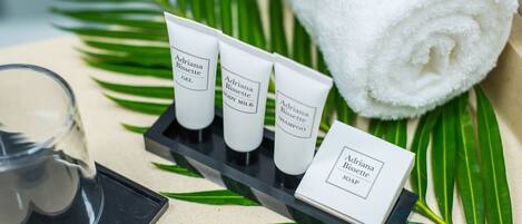 A tray with three tubes of Adriana Bissette body care products, a rolled white towel, and a glass of water on a surface with green leaves.