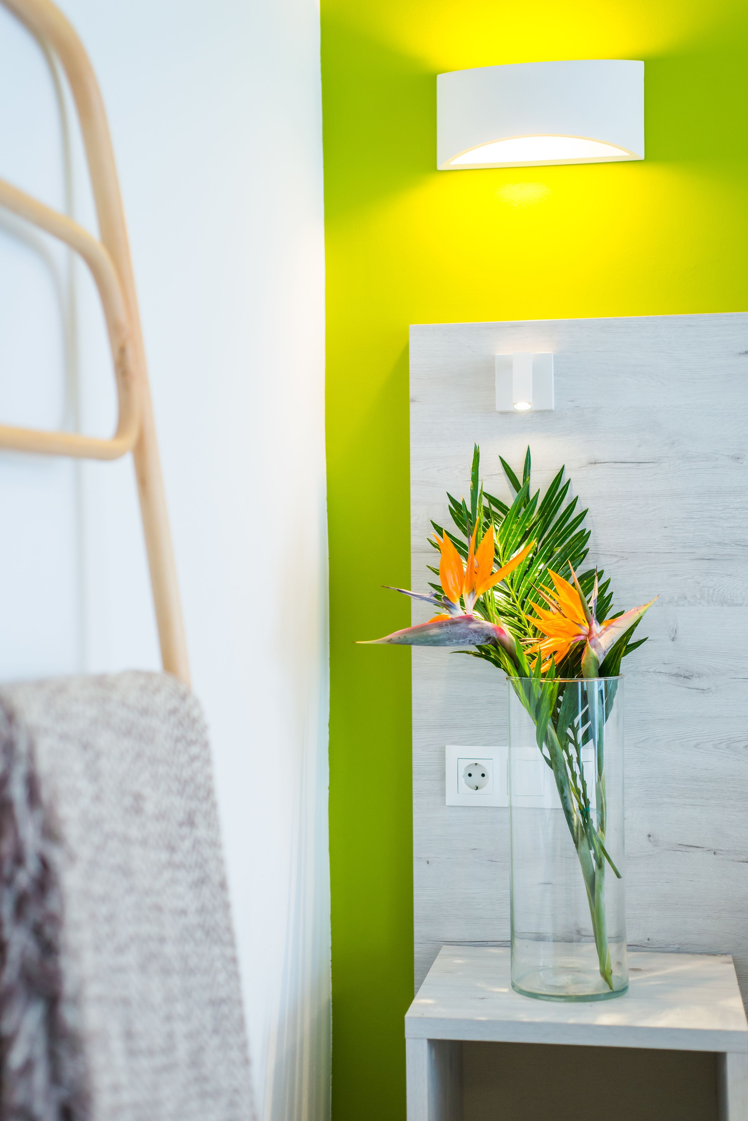 Un baño con una toalla blanca colgada en un perchero, un jarrón de vidrio con flores sobre un estante blanco, y una lámpara de pared.