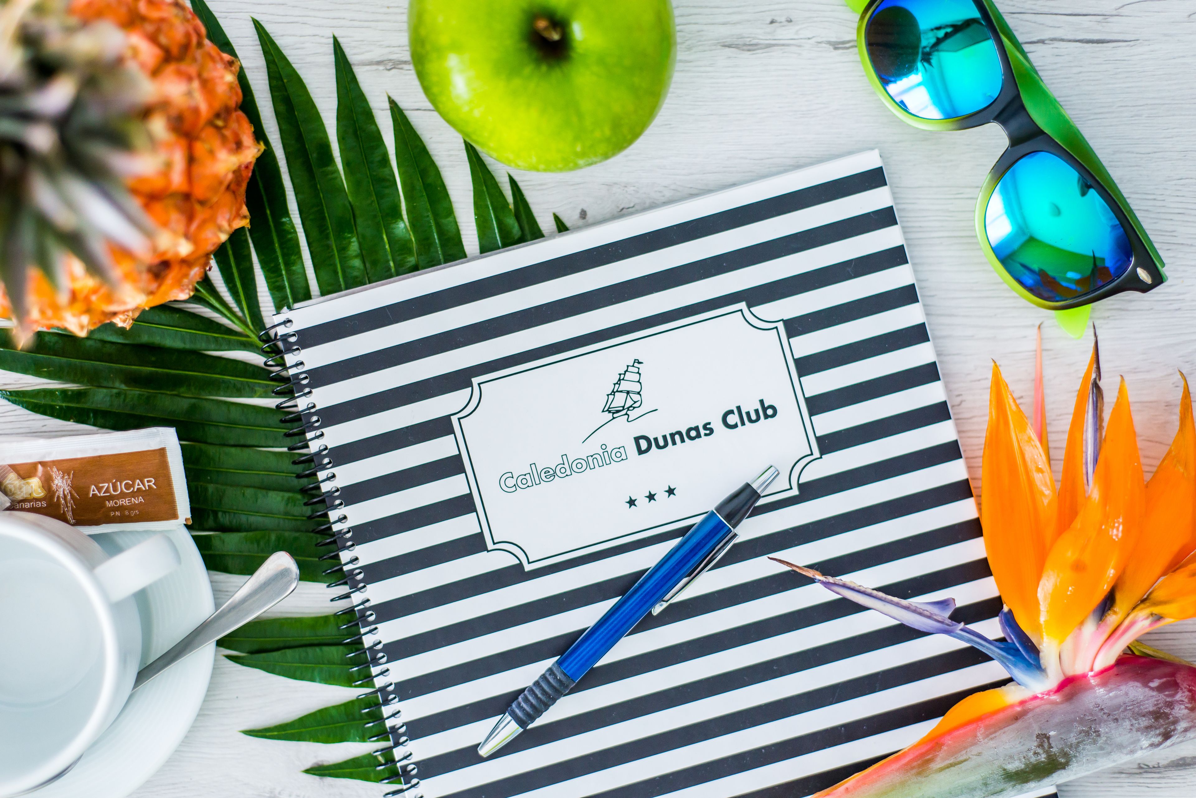 Un cuaderno con un logotipo de equipo, un bolígrafo, una piña, una manzana, gafas de sol y una flor colorida.
