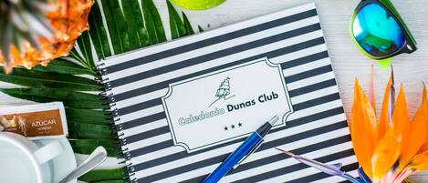 Un carnet orné du logo d’un club, un stylo, un ananas, une pomme, des lunettes de soleil et une fleur aux couleurs vives.