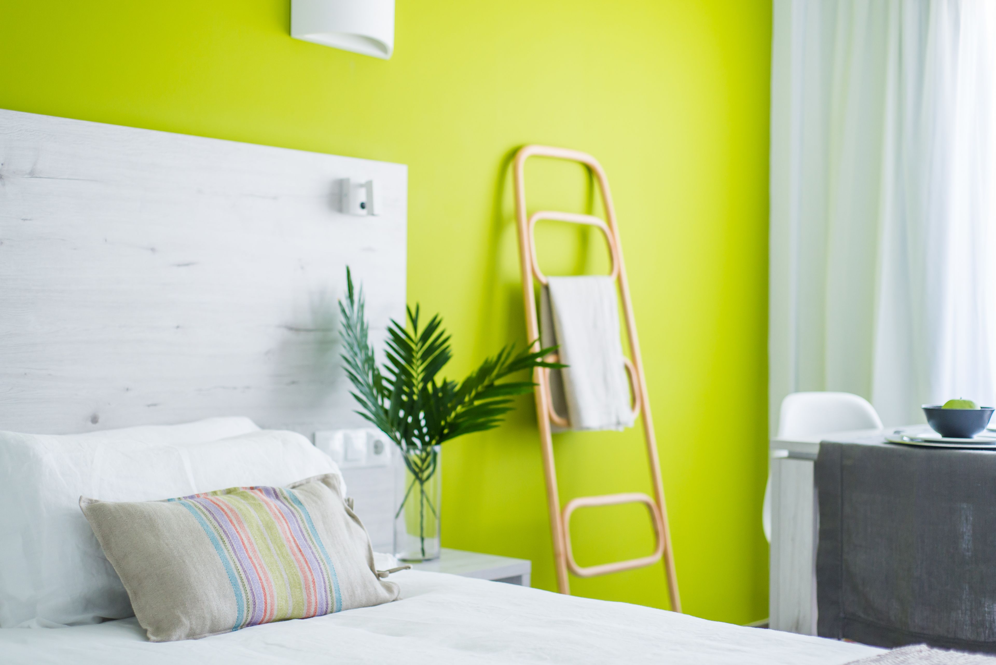 A hotel room with a white bed, a colorful striped pillow, a small potted plant, and a wooden ladder against a bright green wall.