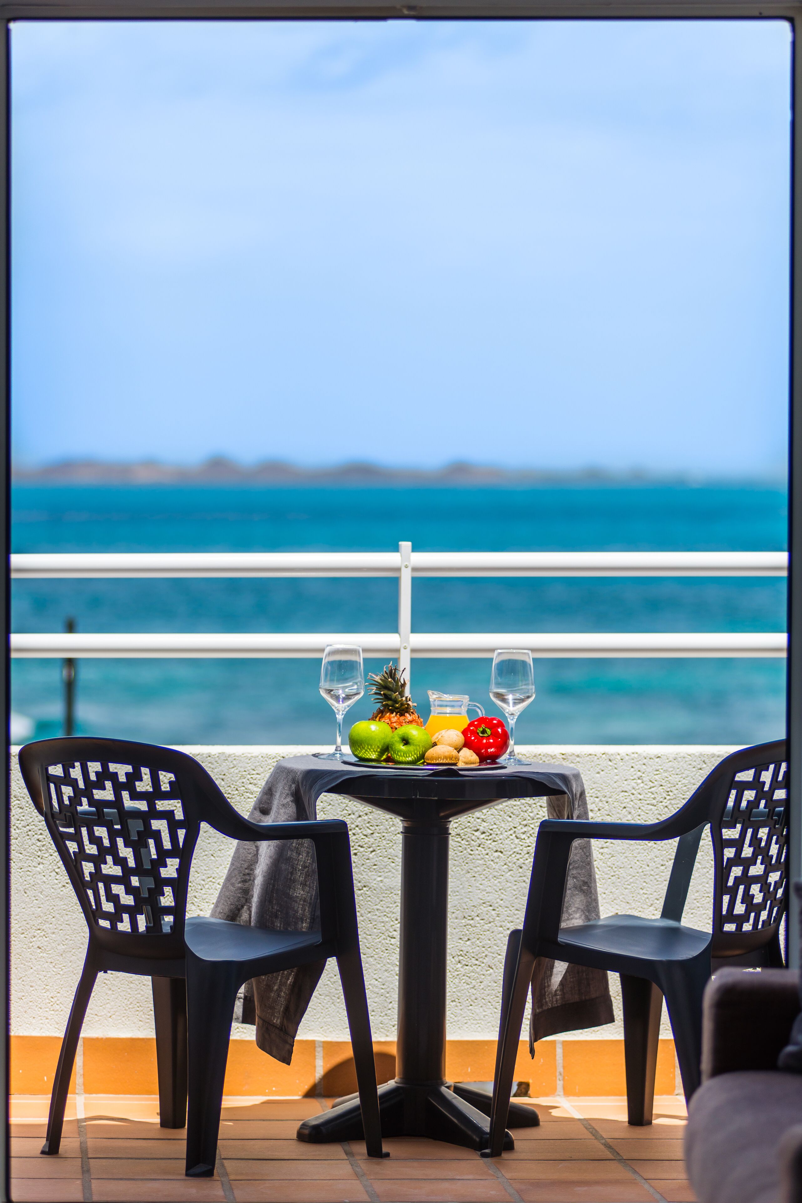 Una mesa con un frutero, dos copas de vino y una jarrón en un balcón con vistas al mar.