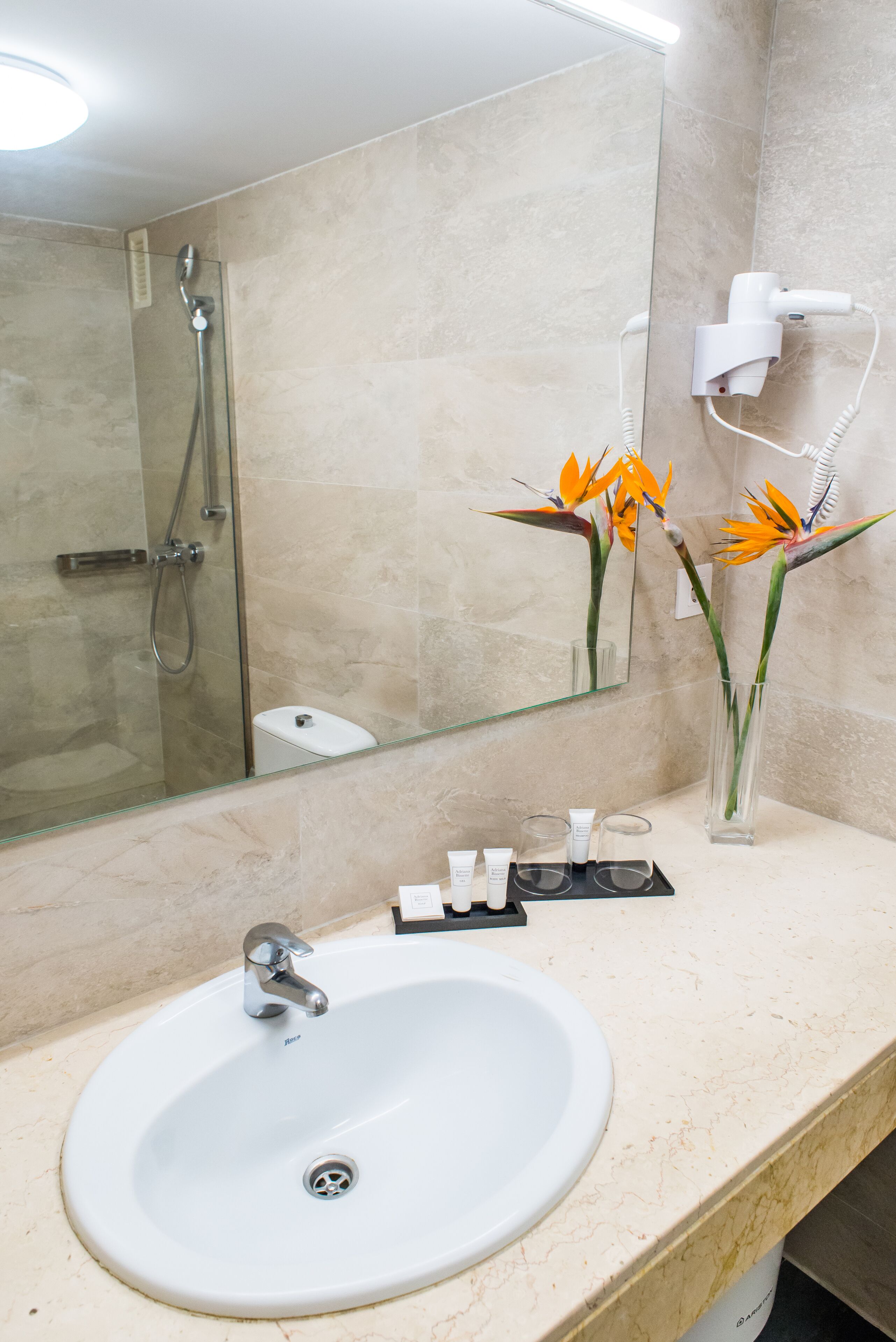 A bathroom with a white sink, a glass shower enclosure, a mirror, and a vase with flowers.