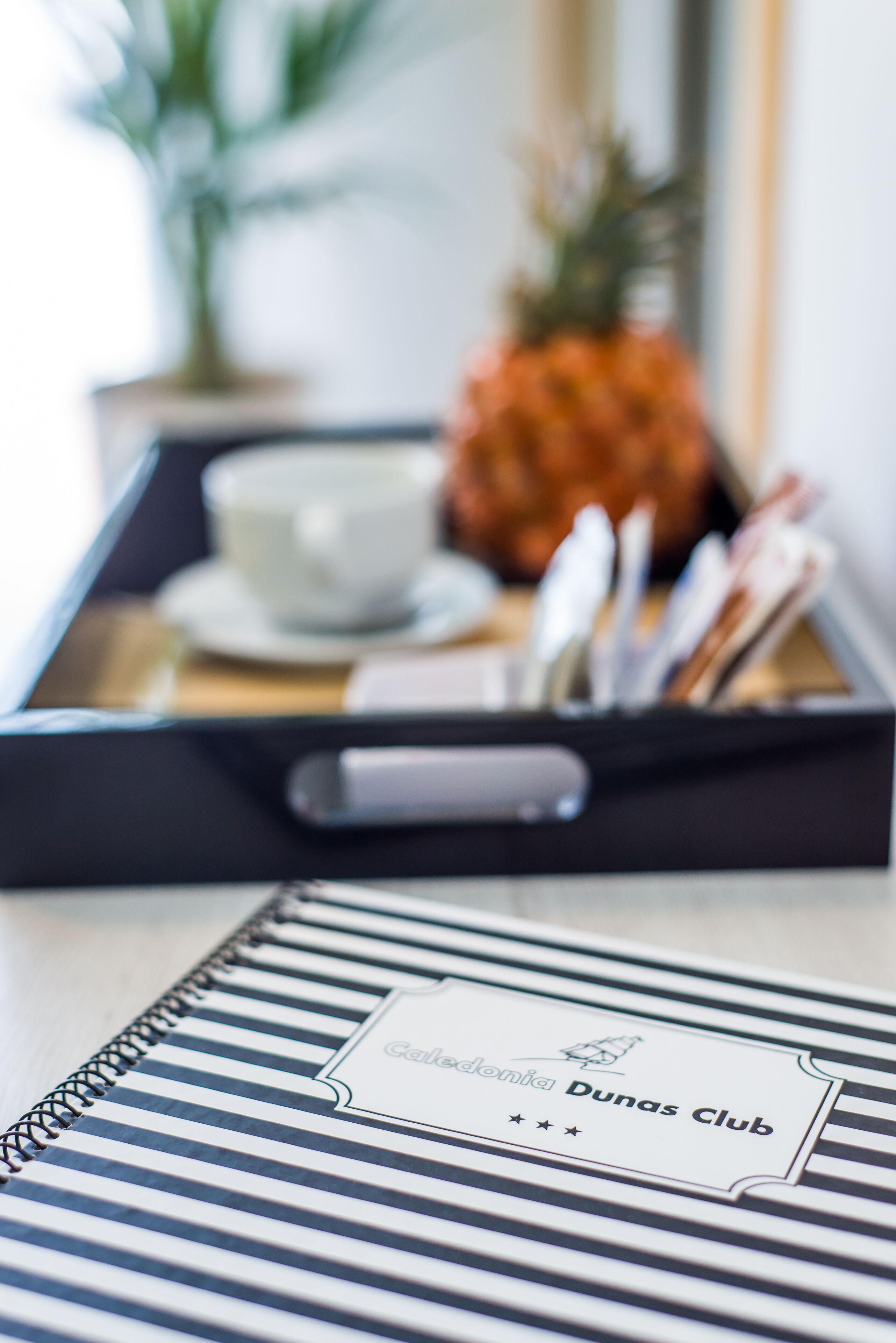 Una bandeja con una taza, una cuchara y una piña, además de un cuaderno con el rótulo 'Dunas Club'.
