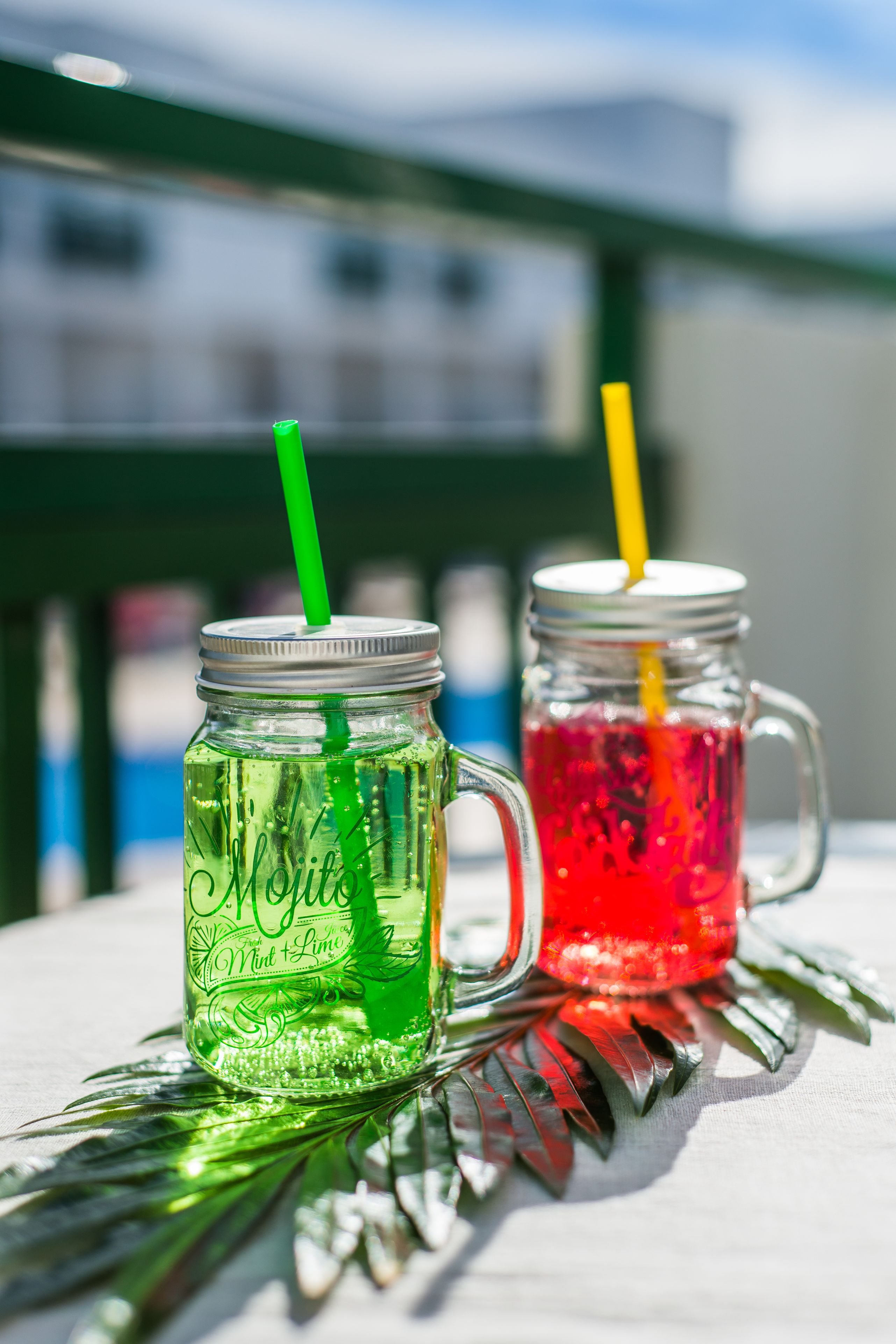 Two glass jars with straws, one labeled 'Mint' and the other 'Lime,' placed on a table with a green railing in the background.