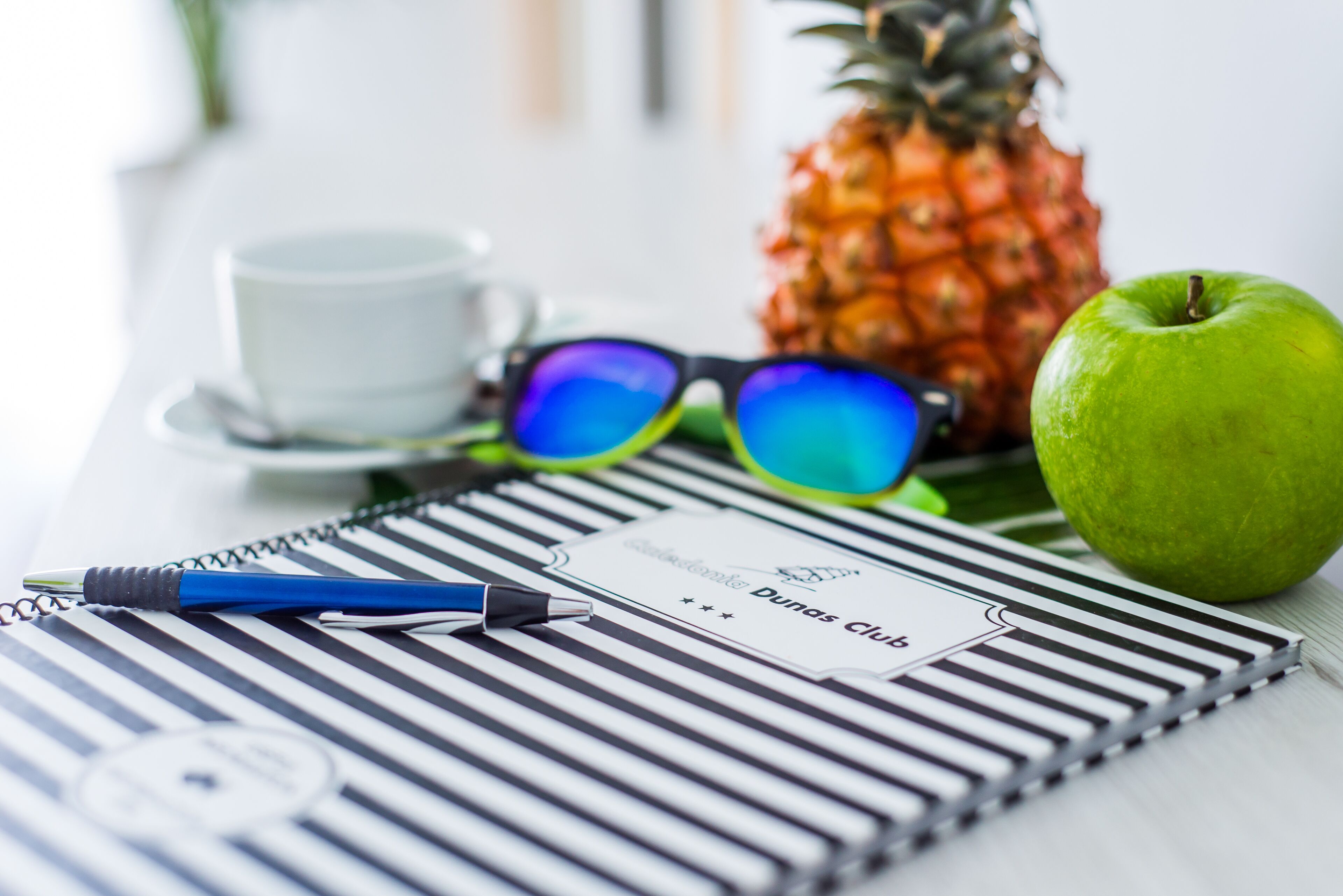 A notebook with a business card, a pen, a pineapple, an apple, and a cup on a table.
