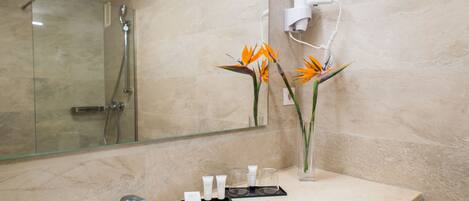 A bathroom with a glass shower enclosure, a sink with toiletries, and a hairdryer on the wall.